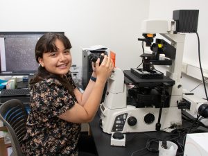 Summer research program student Elaine Torres-Lopez sits in front of a microscope, looking at the camera and smiling.
