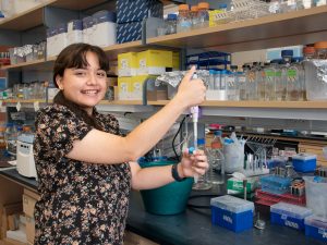 Summer research program student Elaine Torres-Lopez stands next to a lab bench, holding a pipette and smiling.