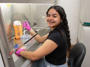 Gabriela P. Romero-González, a summer research program student, smiles at the camera while holding a pipette under a lab hood. 