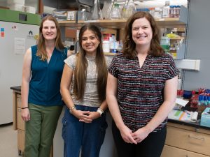 Noah Venable, a summer research program student, stands next to her mentor, Catherine Pettinger, and PI, Erica Majumder, in front of a lab bench. All three are smiling at the camera. 