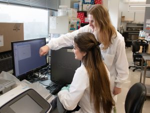 Noah Venable, a summer research program student, sits in front of a computer while her mentor, Catherine Pettinger, stands next to her, leaning over the computer to point at the screen. 
