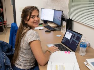 Noah Venable, a summer research program student, turns toward the camera and smiles while seated in front of a computer. 