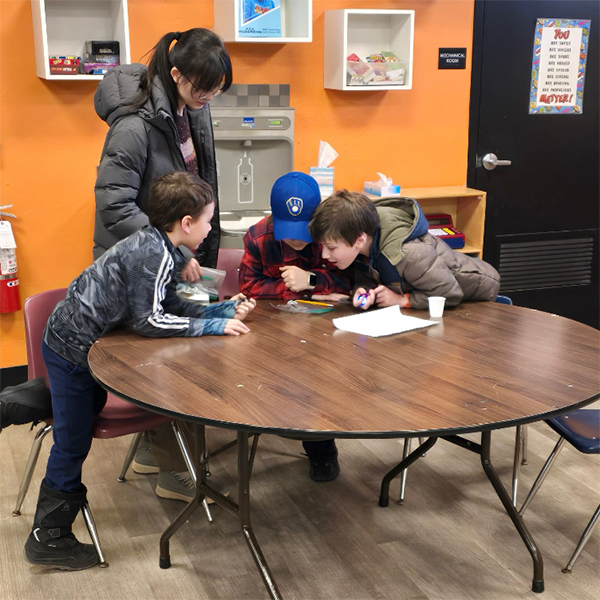 A student from Service with Youth in STEM (INTEGSCI 240) stands at a table helping three elementary school students with a project.