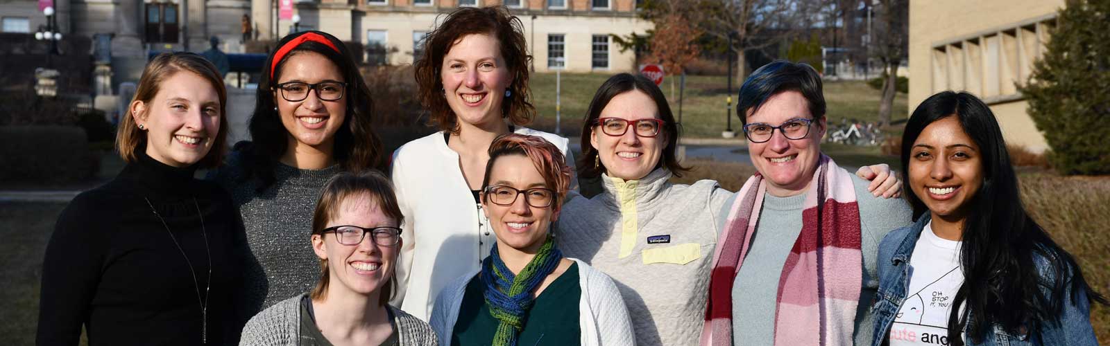A professor and seven graduate students pose together outside. 
