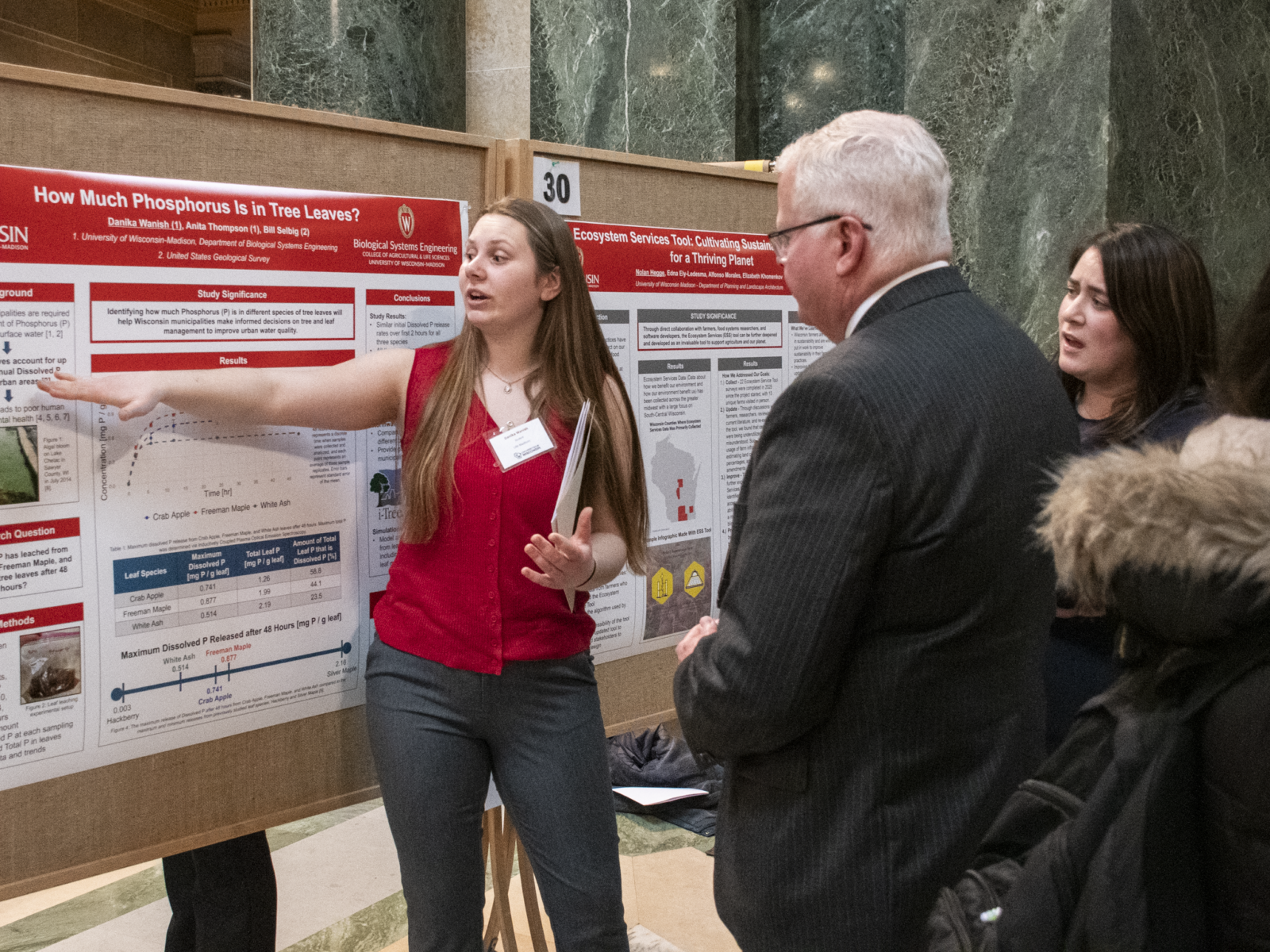 A student researcher presents her poster at Research in the Rotunda. She is pointing at her poster and talking while an attendee listens to her.