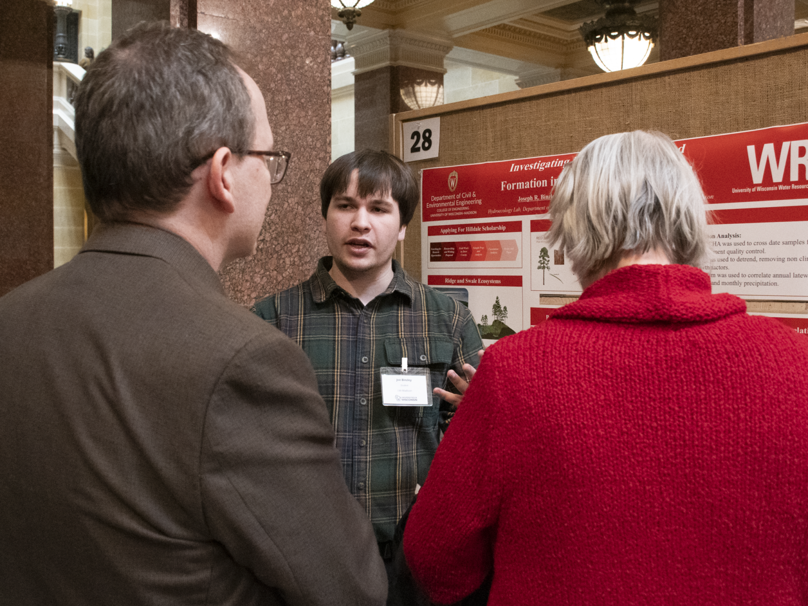A student researcher presents his poster at Research in the Rotunda, while two attendees with their backs facing the camera listen to him.