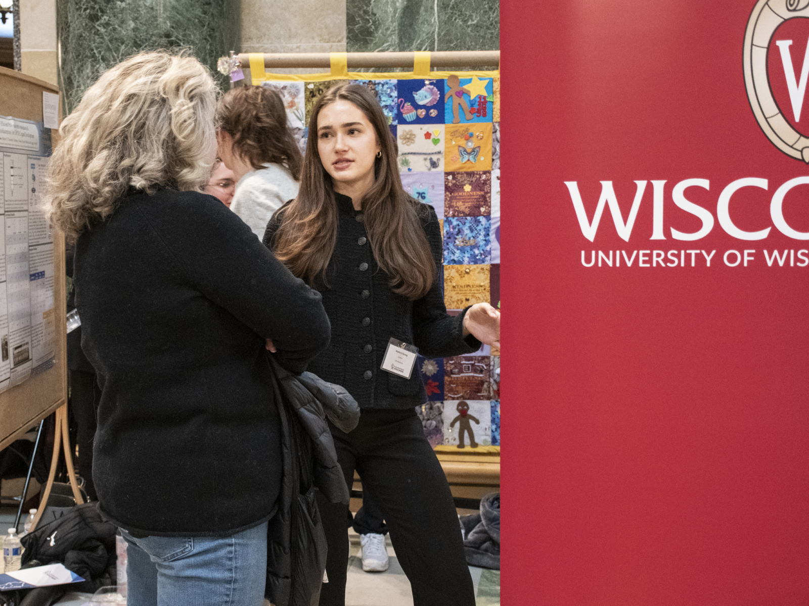 A student researcher presents her poster at Research in the Rotunda. She is gesturing at her poster and facing an attendee, who has their back to the camera.