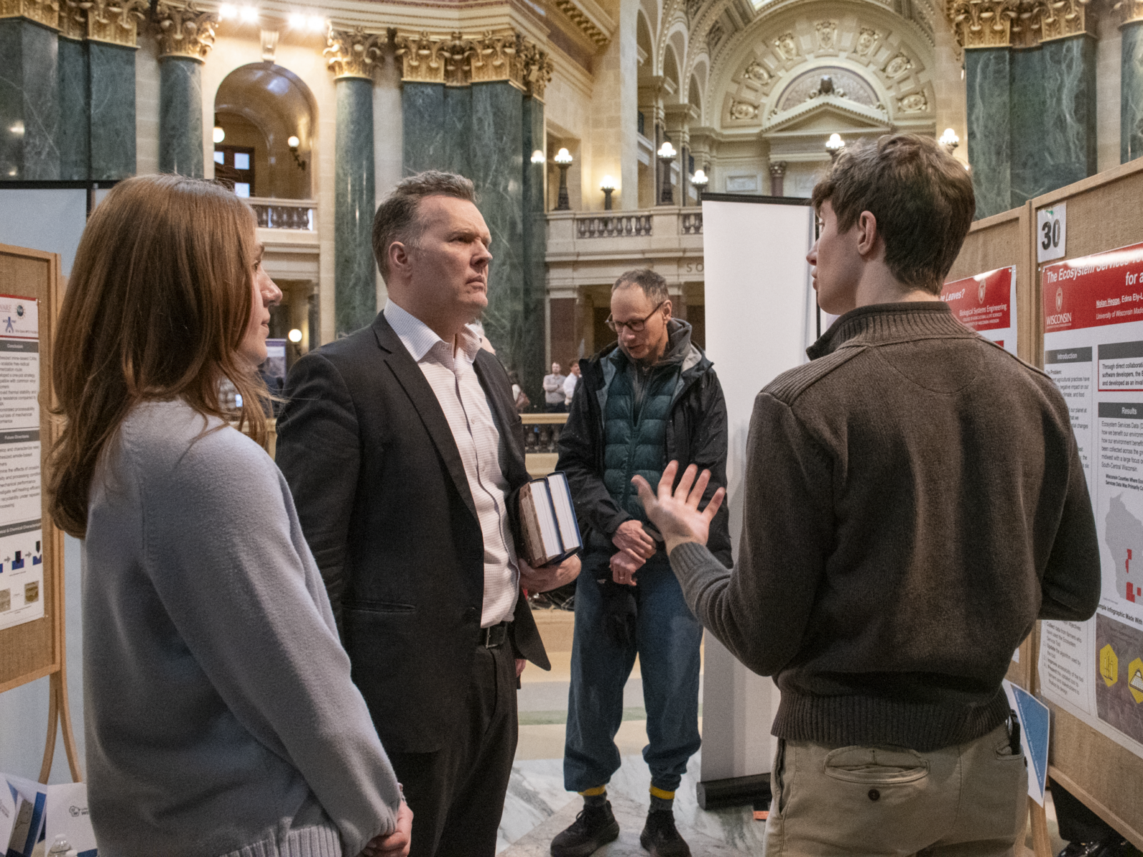 A student researcher presents his poster at Research in the Rotunda. His back is to the camera, but we can see the side of his face as he talks to two attendees.