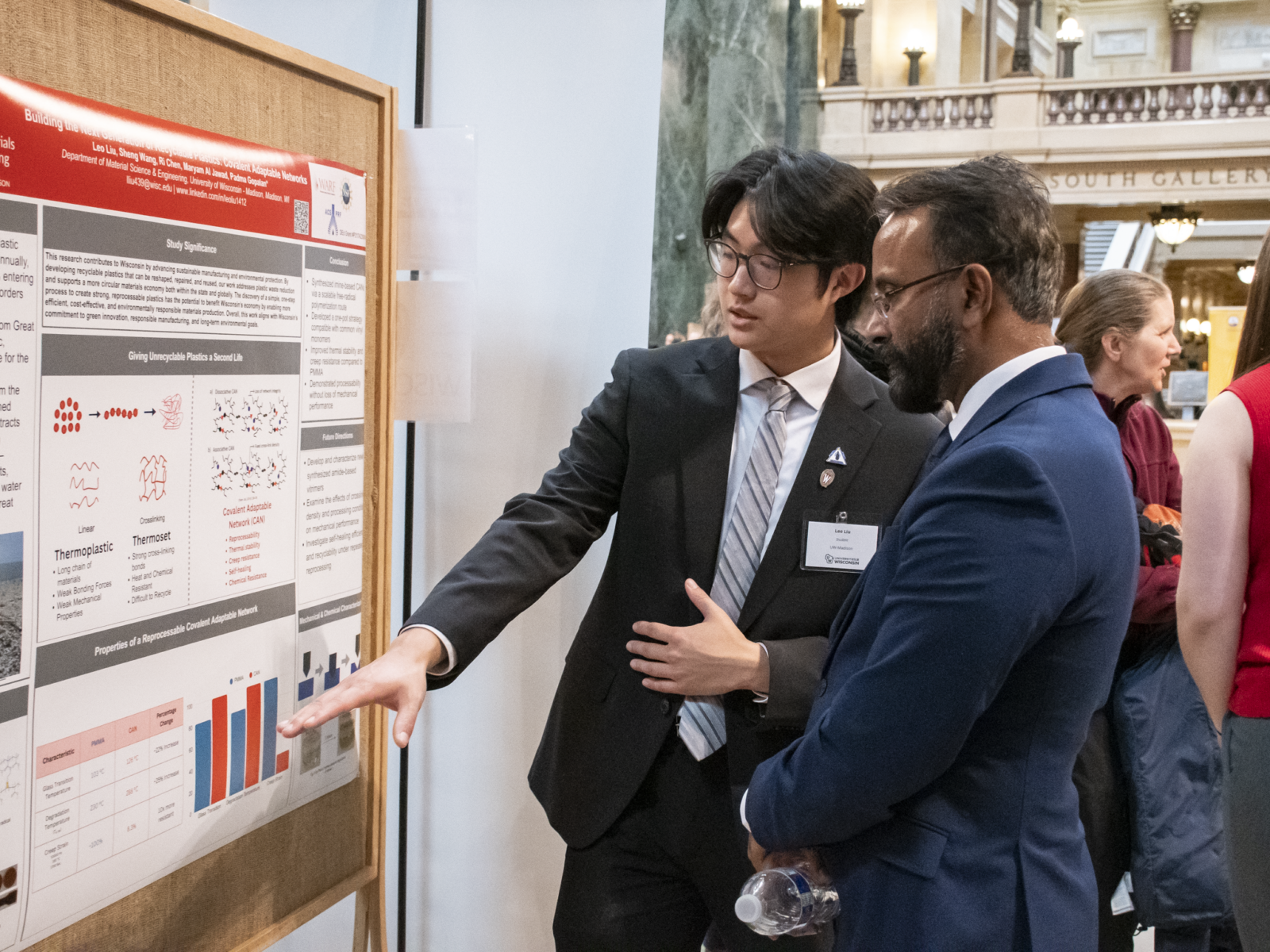 A student researcher presents his poster at Research in the Rotunda. He is pointing to his poster and talking while an attendee looks on.