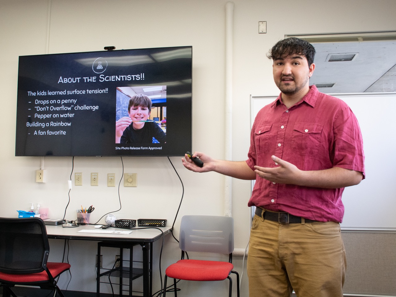 A student gives a presentation while standing to the side of a large screen, gesturing towards it. It reads "About the Scientists!!" and has a photo of an elementary school student doing a science experiment.