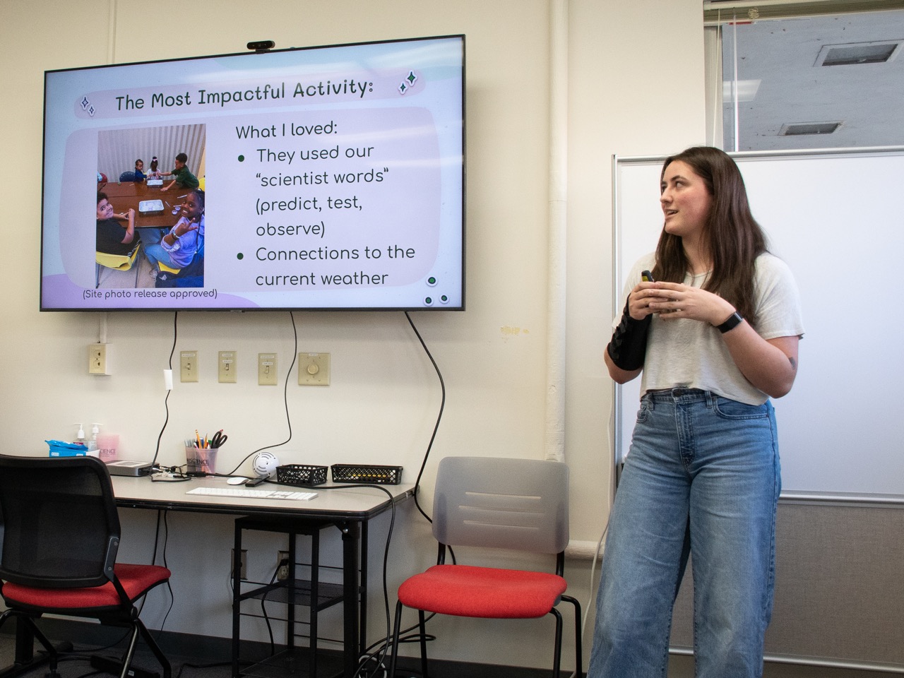 A student gives a presentation while standing to the side of a large screen, which reads, "The Most Impactful Activity" and has a photo of elementary school students doing a science experiment.