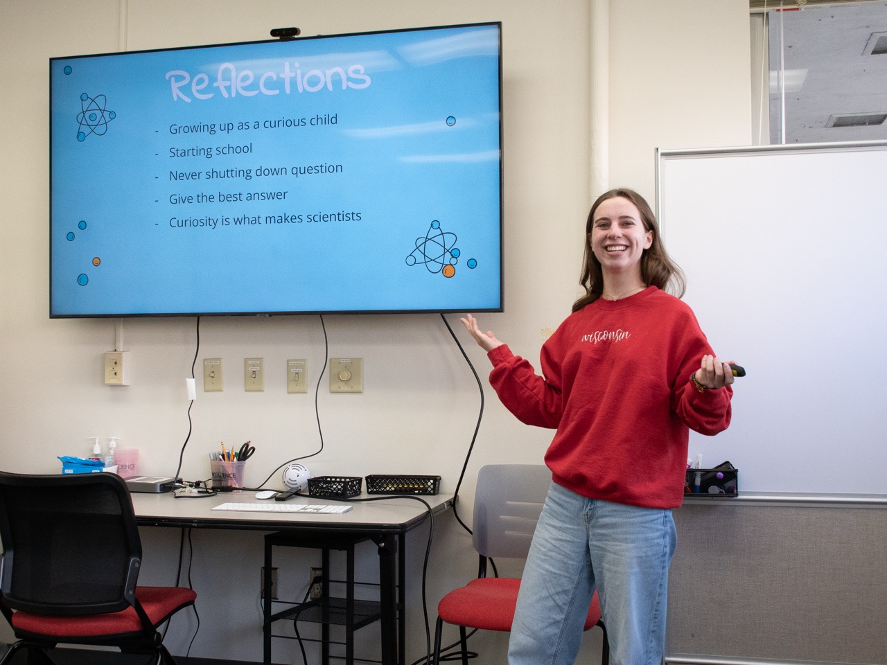 A student smiles and gestures at a screen that reads "Reflections."