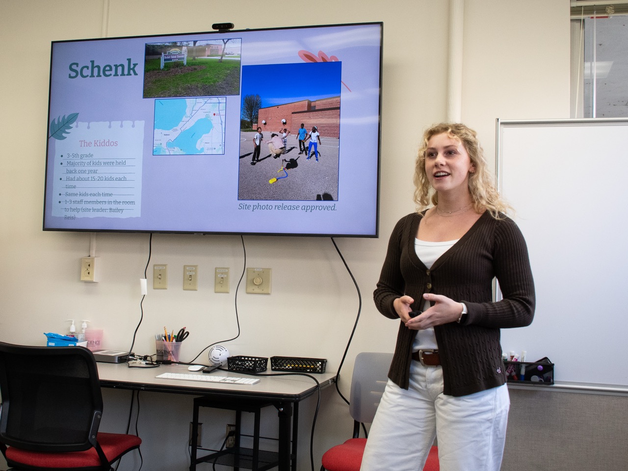 A student gives a presentation while standing to the side of a large screen, which has the word "Schenk," a map, and a photo of kids doing a science activity.
