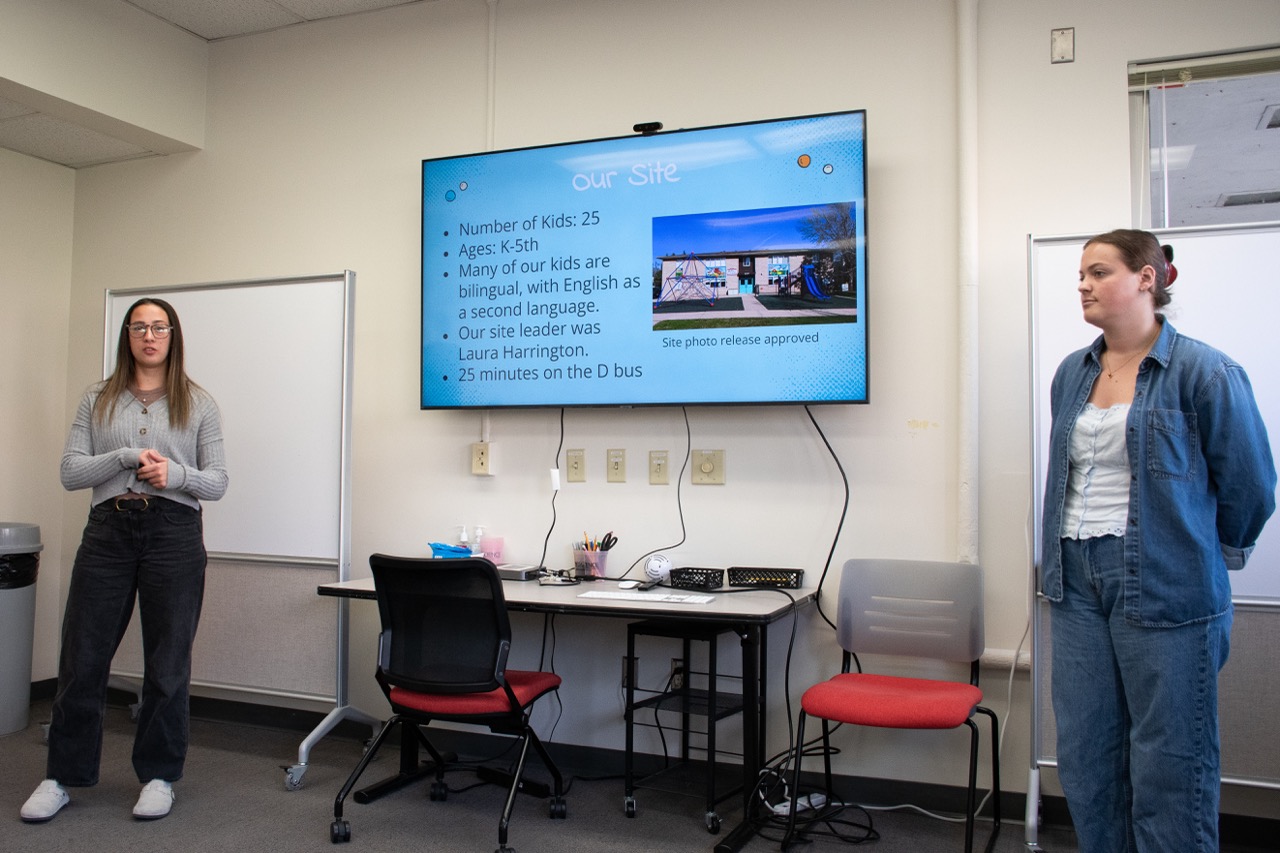 Two students give a presentation standing next to a large screen that reads "Our Site" and has a photo of a school building.