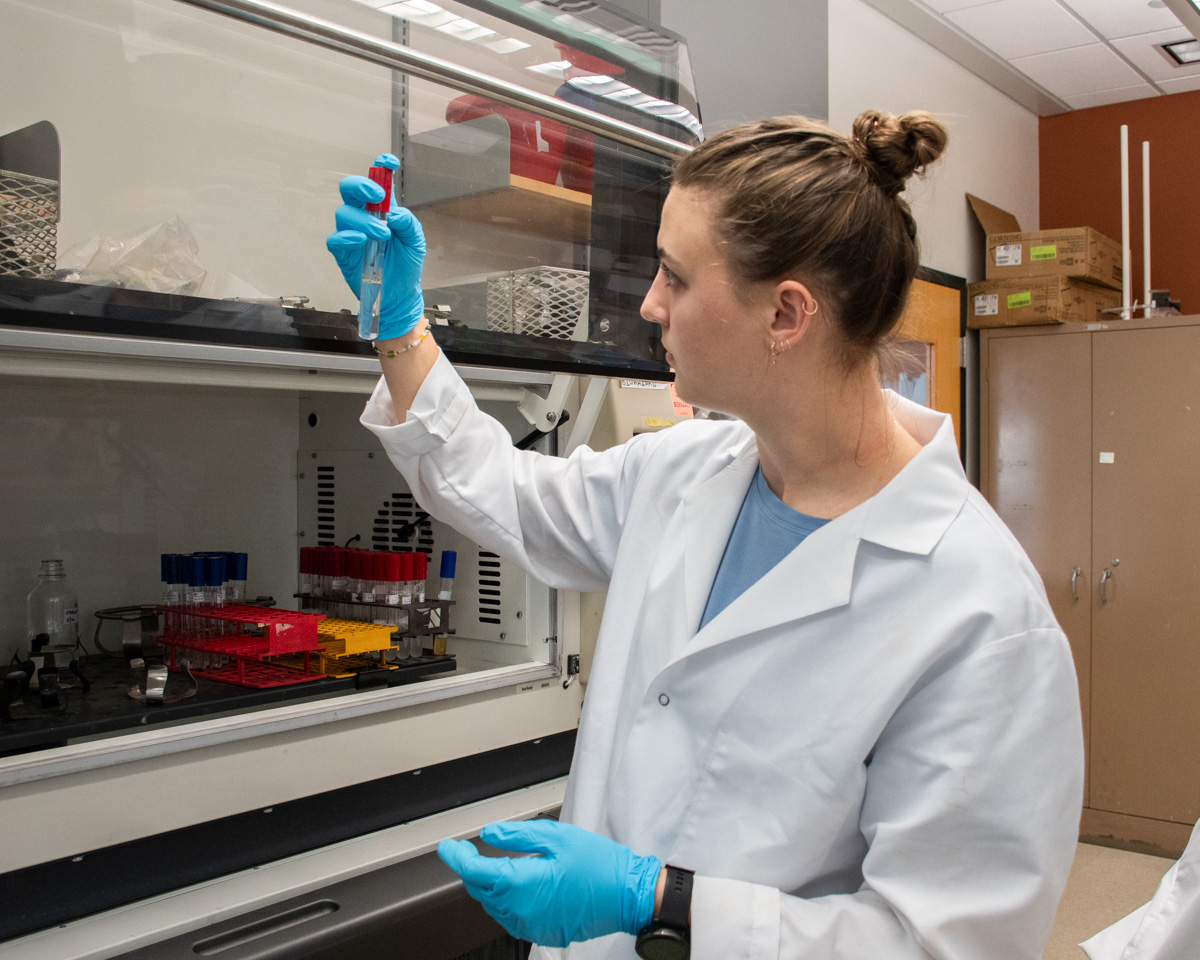 A student stands in a lab wearing a white lab coat and blue gloves. She is holding up a test tube and looking at it.