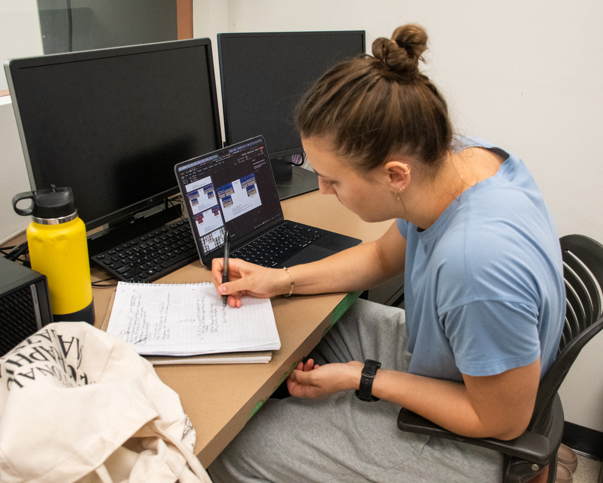 A student sits at a desk in front of an open laptop and two computer screens. She is writing in a notebook.