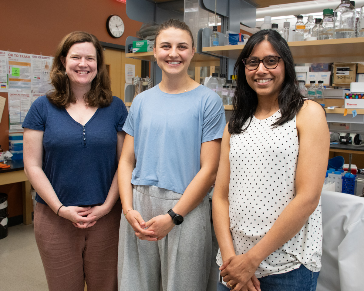 Three women are smiling as they pose together in front of a lab bench.