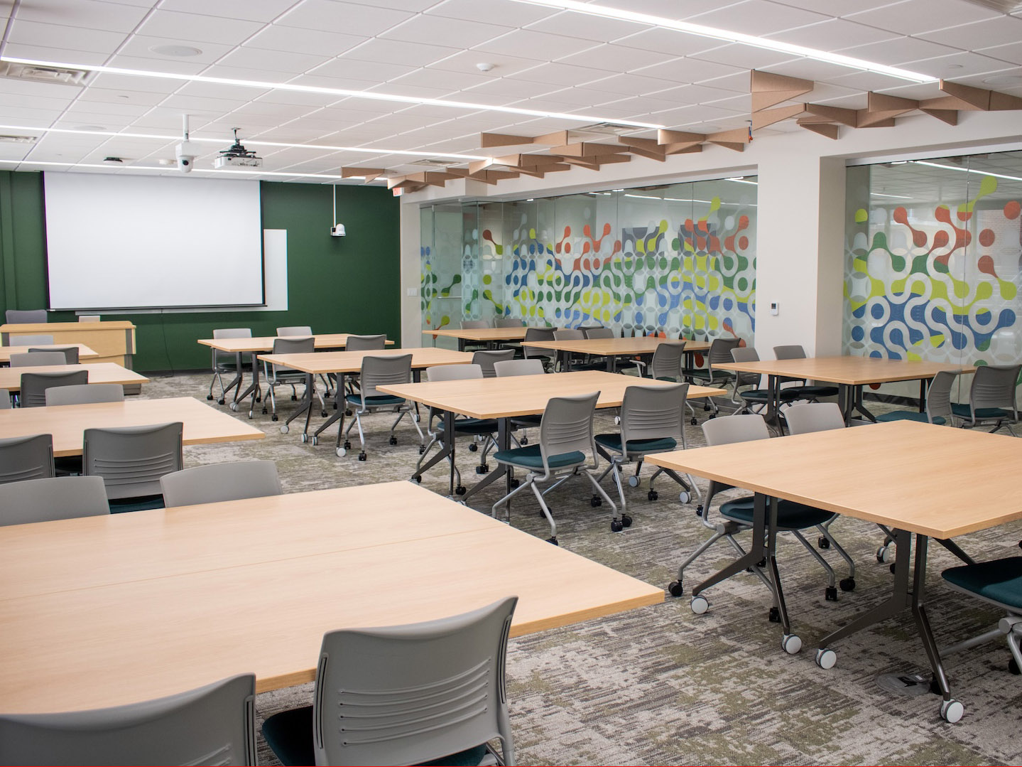 A large classroom with about a dozen rectangular tables with chairs. There is a projection screen at the front, and a glass wall with colorful diagrams on the right.