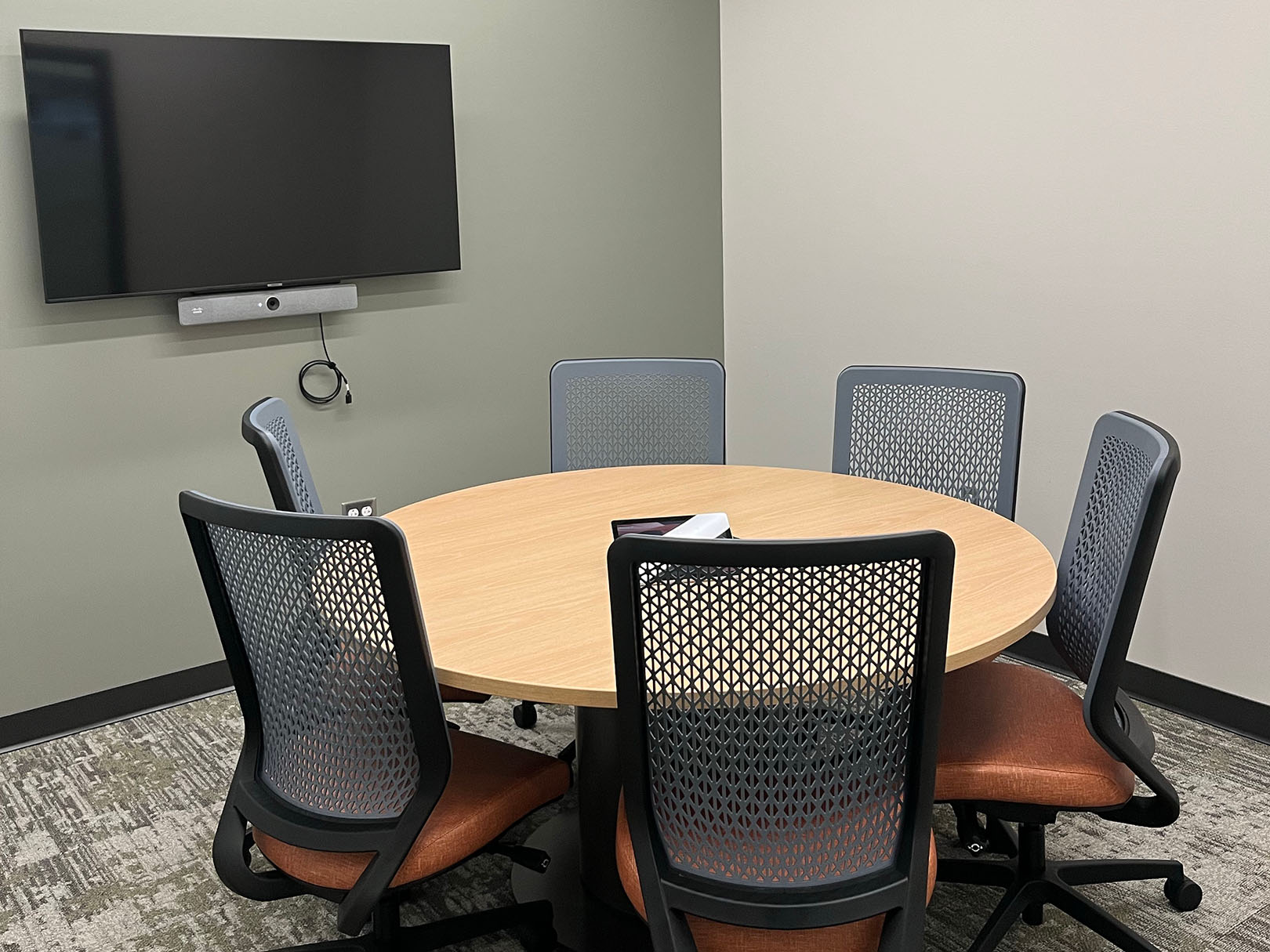 A room with a round table, several desk chairs, and a wall-mounted TV screen.