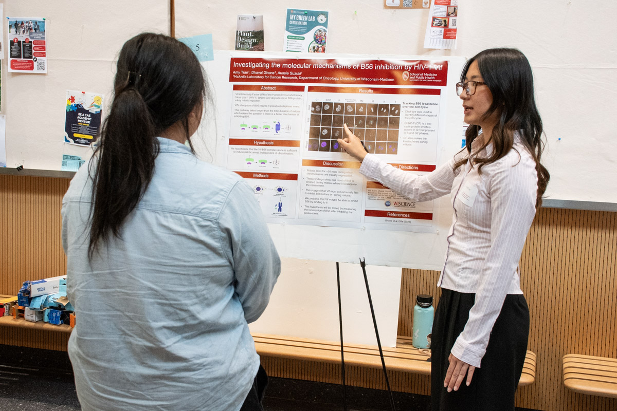A student points at her research poster poster while talking to an onlooker at during a poster session.