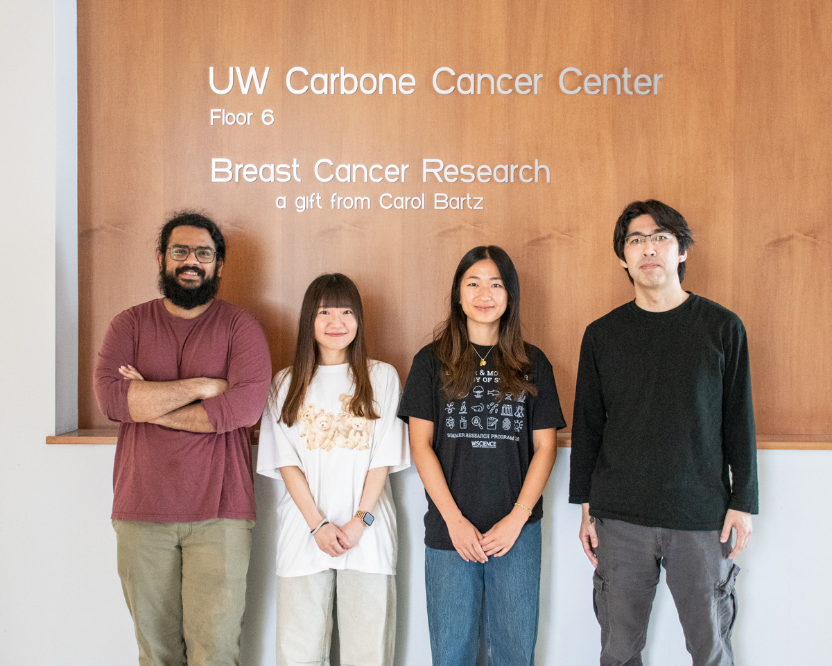 Four lab members pose together in front of a wall that reads, "UW Carbone Cancer Center, Floor 6. Breast Cancer Research, a gift from Carol Bartz."