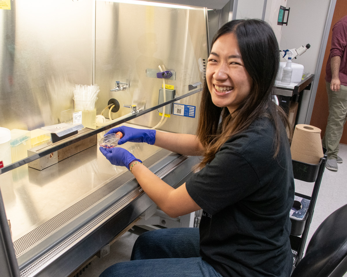 A student wearing blue gloves holds a container under a hood in a lab. Her face is turned towards the camera with a wide smile.