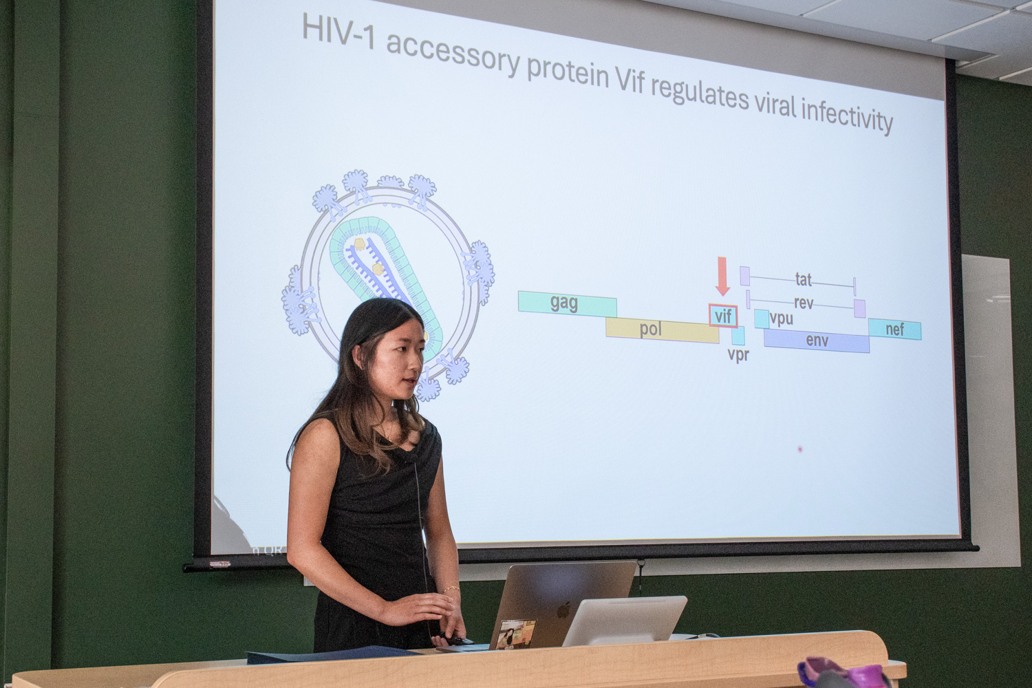 A student in a black sleeveless shirt stands and talks in front of a projector screen. The screen reads, "HIV-1 accessory protein Vif regulates viral infectivity," and shows a science diagram.