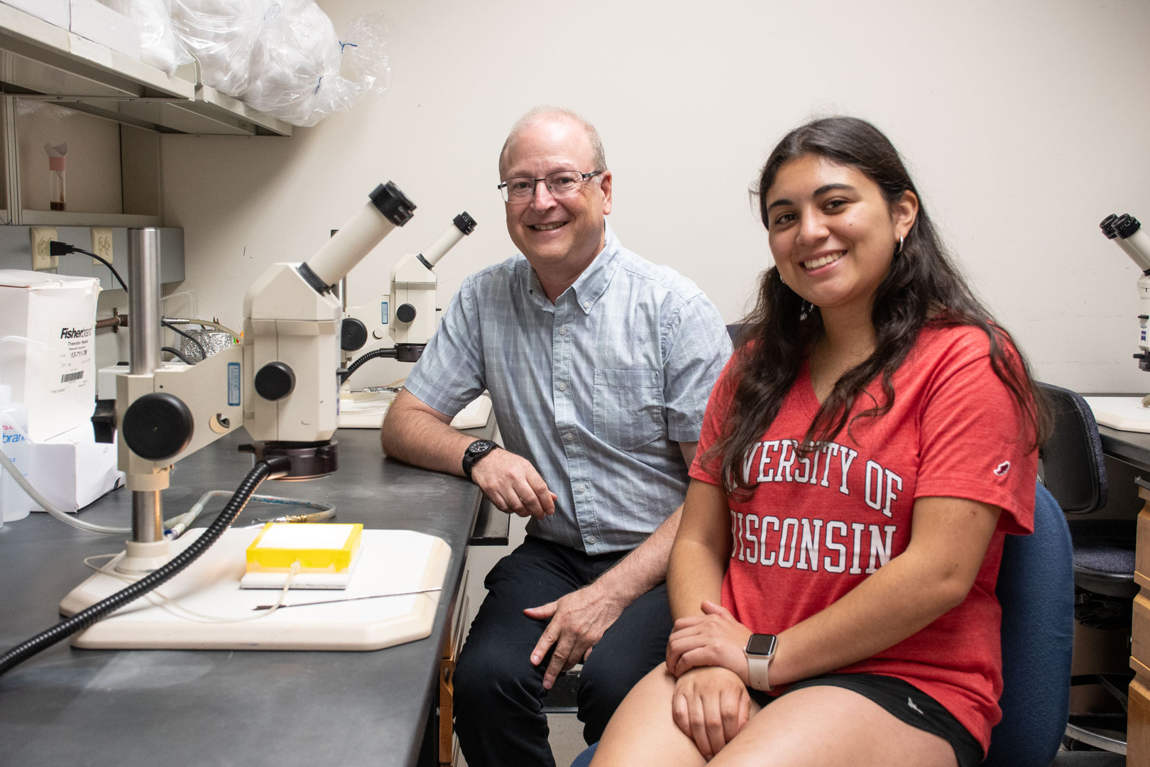 A student and a professor are smiling as they next to each other at a lab bench with microscopes.