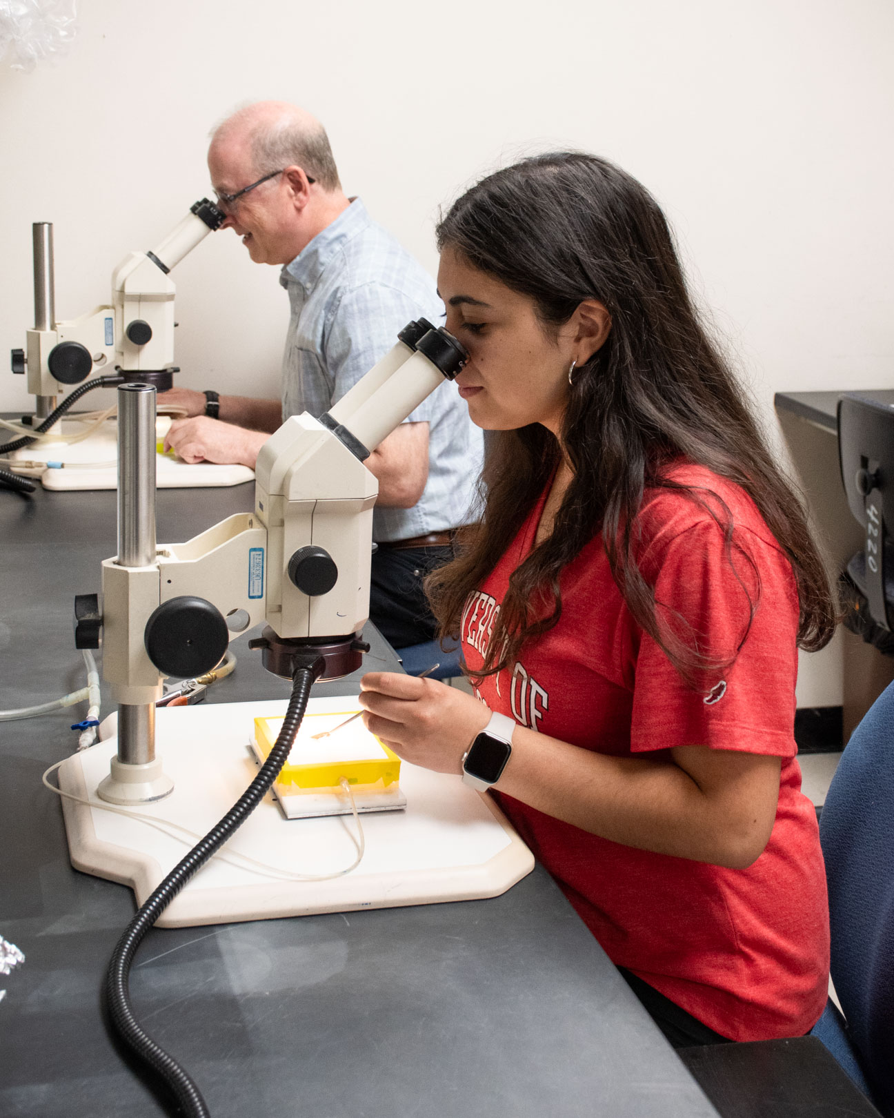 A student and a professor sit next to each other at a lab bench and look into microscopes.