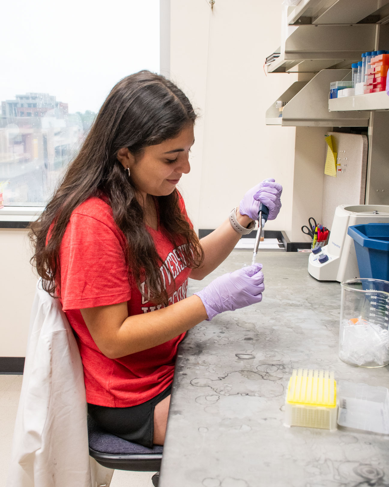 A student wearing a red shirt and purple gloves uses a pipette in a lab.