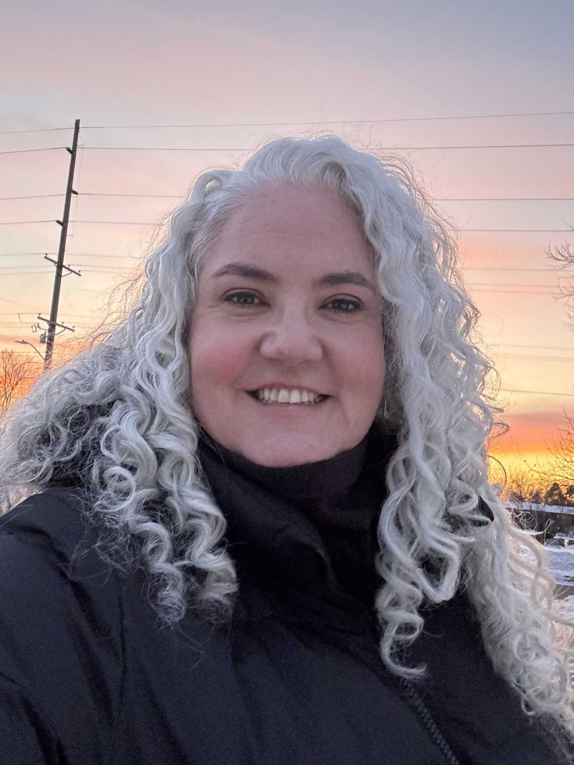 A woman with shoulder-length curly silver hair wearing a black coat. There is a sunset in the background.