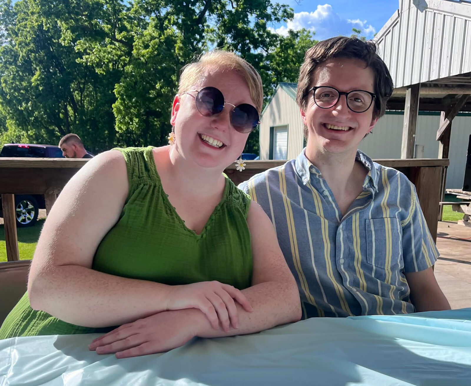 Two people sit next to each other smiling at an outdoor picnic table. One is a woman wearing a green dress and sunglasses, and the other is a man wearing a striped shirt and glasses.