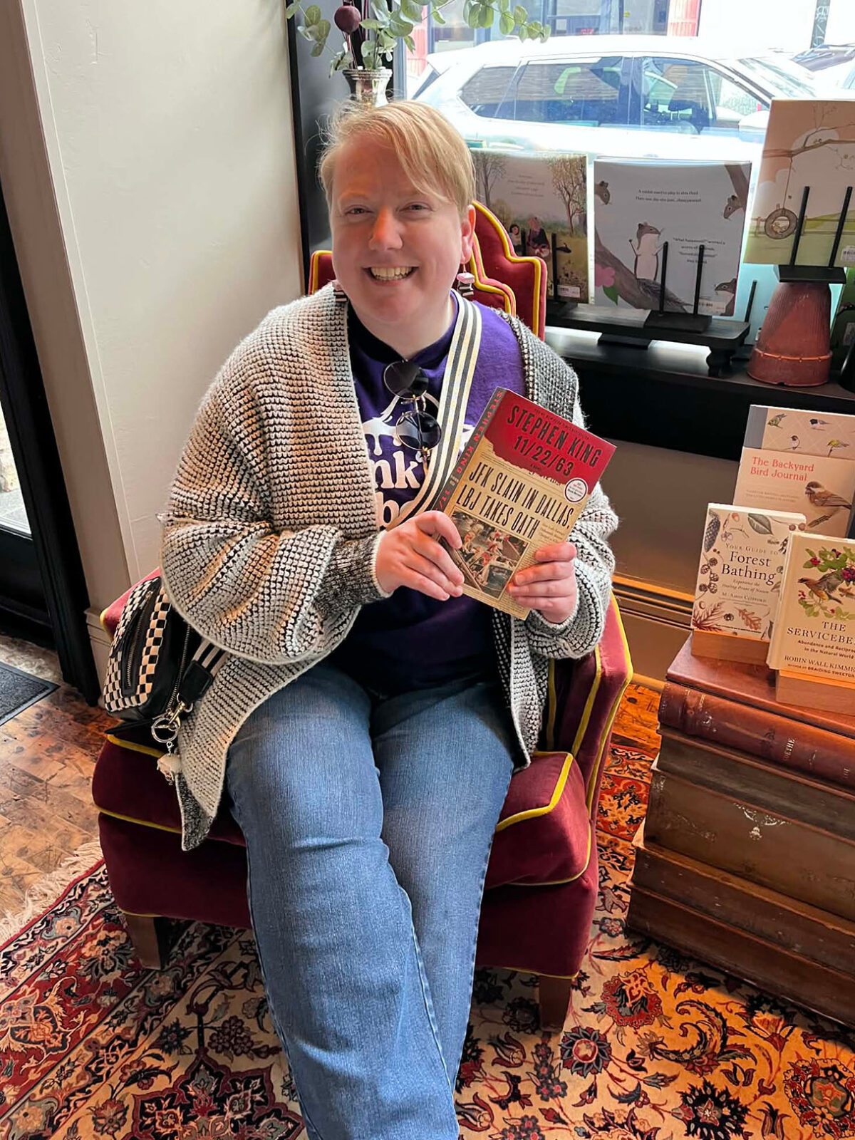 A smiling woman wearing a gray and white sweater sits on a chair in a bookstore while holding up the book 11/22/63 by Stephen King.