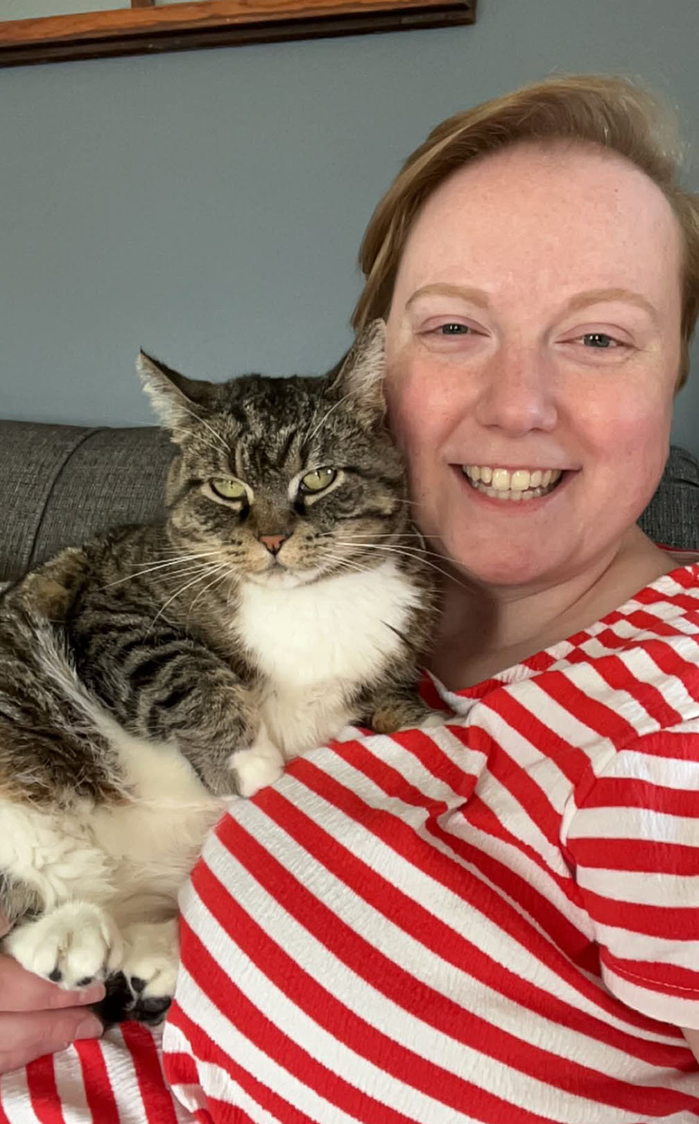 A smiling woman wearing a red and white striped shirt sits on a couch with a cat cuddled up to her face.